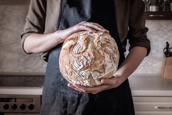 Mulher de avental preto no fundo da cozinha com mãos a segurar um pão redondo